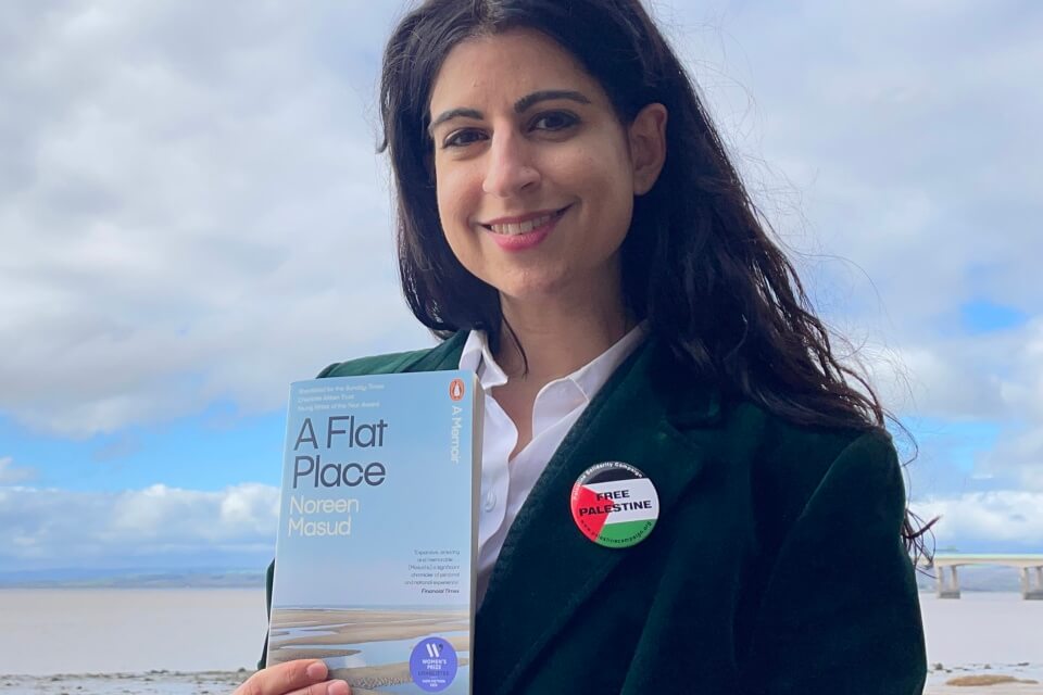 Colour photographic portrait of Noreen Masud stood on a sandy plain holding her book